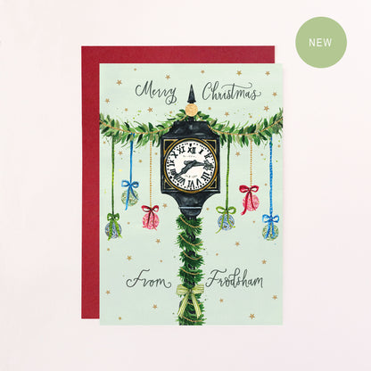 A festive greeting card with green calligraphy text reading 'Merry Christmas From Frodsham' with a drawing of Frodsham Town Clock, baubles, bows and faux glitter stars.