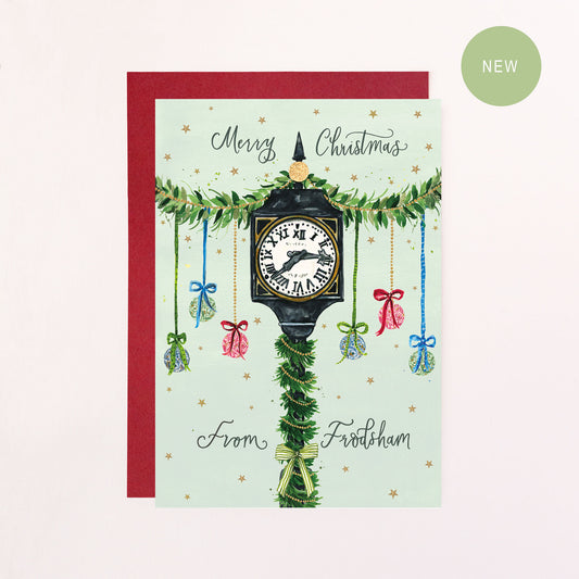 A festive greeting card with green calligraphy text reading 'Merry Christmas From Frodsham' with a drawing of Frodsham Town Clock, baubles, bows and faux glitter stars.